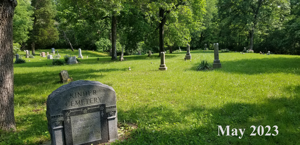 The granite Kinder Cemetery monument standing in the foreground in late spring, with older headstones beyond it on mown green grass beneath oak and cedar trees in full leaf.